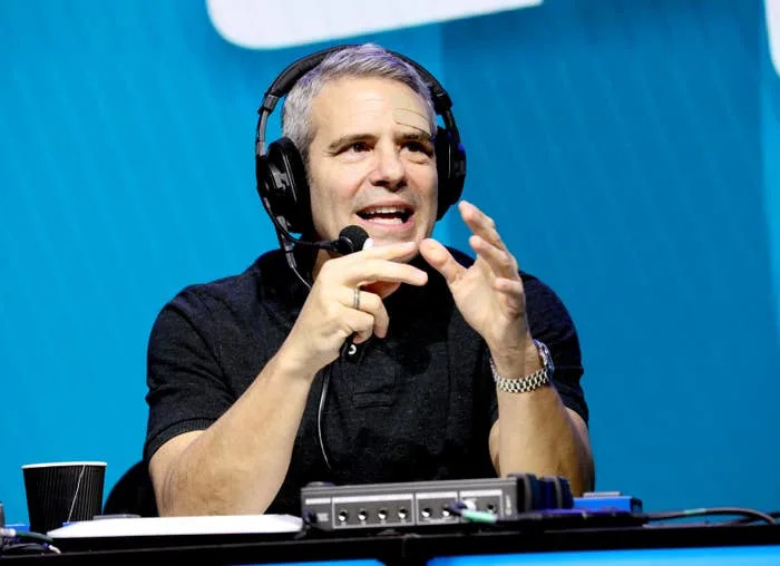 Andy Cohen wearing headphones and a shirt speaks animatedly into a microphone during a broadcast, hands gesturing expressively