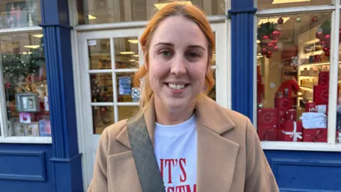 Lisa is smiling. She's wearing a light brown coat and a strap from her handbag is on her shoulder. A shop is behind her.