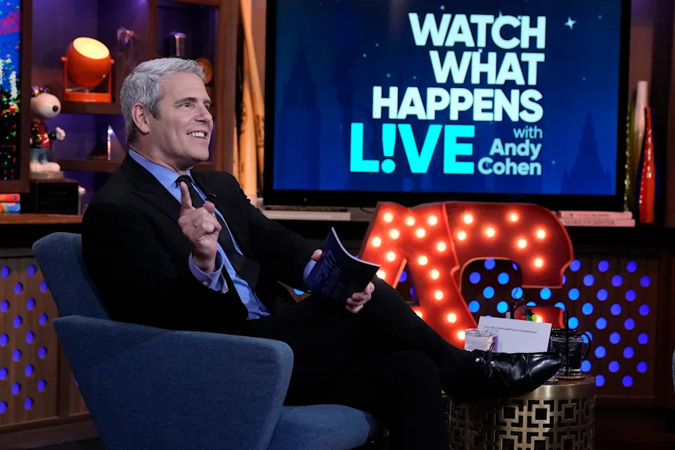 Andy Cohen in a suit gestures while sitting in a TV studio chair, with "Watch What Happens Live" on the screen behind him