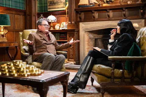 Euan Cherry/BBC Alan Carr, sitting in an armchair, speaking to Claudia Winkleman on the Celebrity Traitors