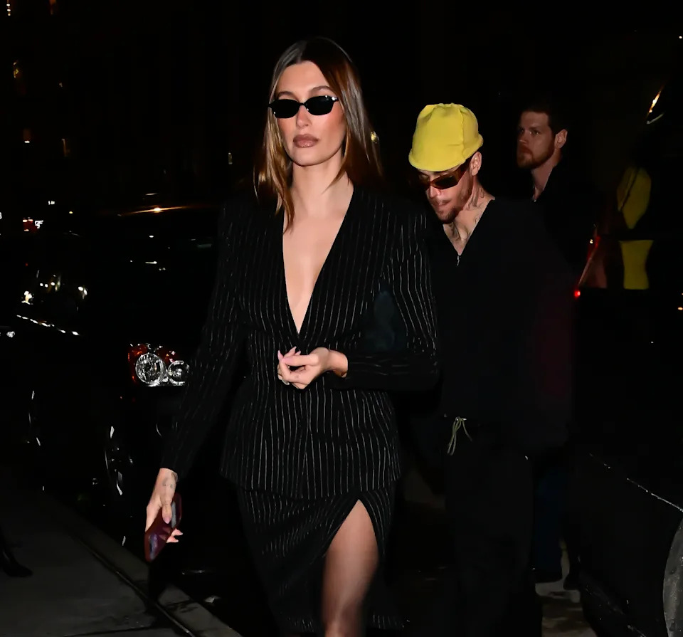 Hailey Bieber in a pinstripe suit with a deep neckline and sunglasses leads, followed by Justin Bieber in a beanie, walking outdoors at night