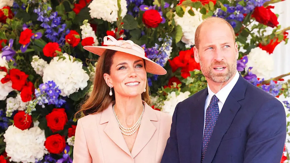 Kaye Middleton smiling and looking up as Prince William looks on as they stand against a wall of blooming flowers.