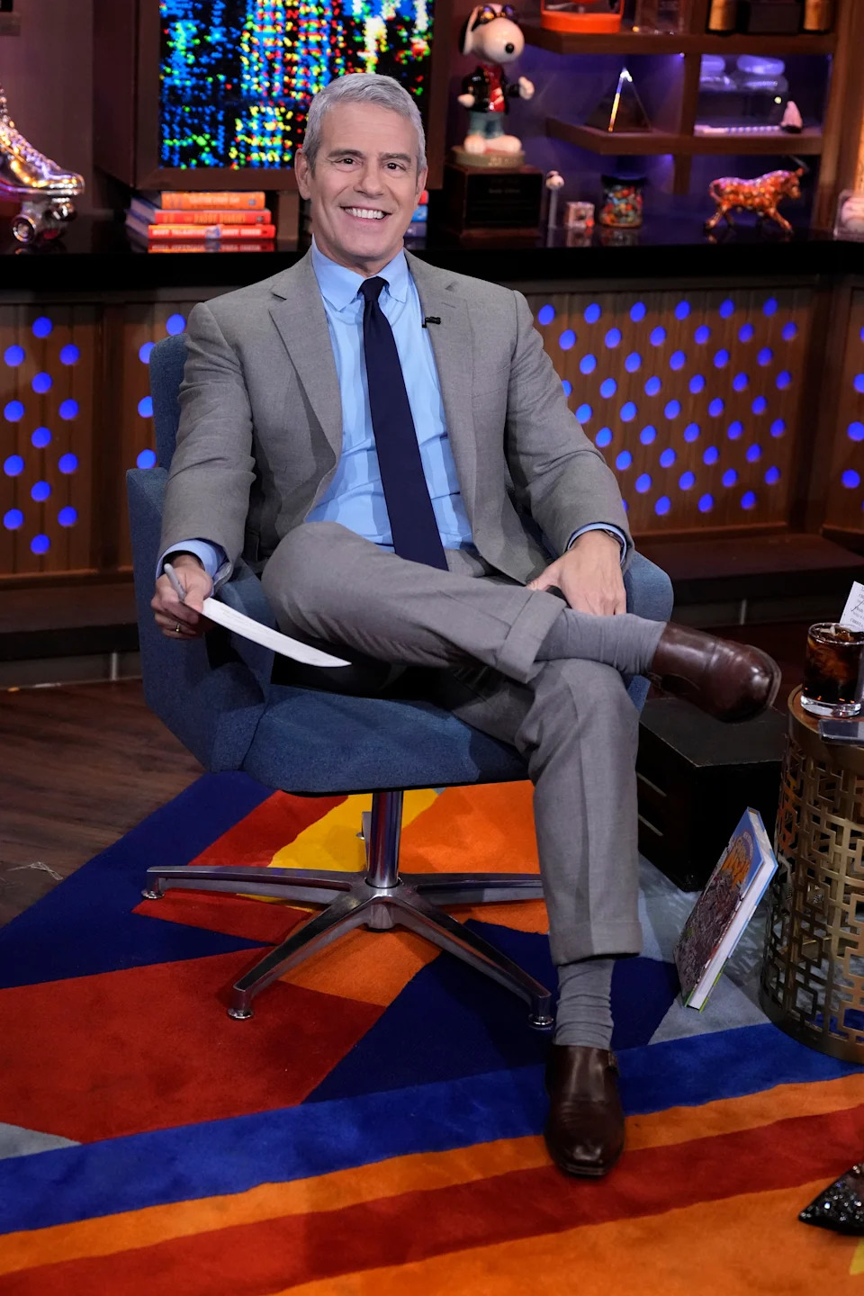Andy Cohen sitting on a chair in a suit with a tie, smiling, on a studio set with colorful decoration and various items on shelves behind
