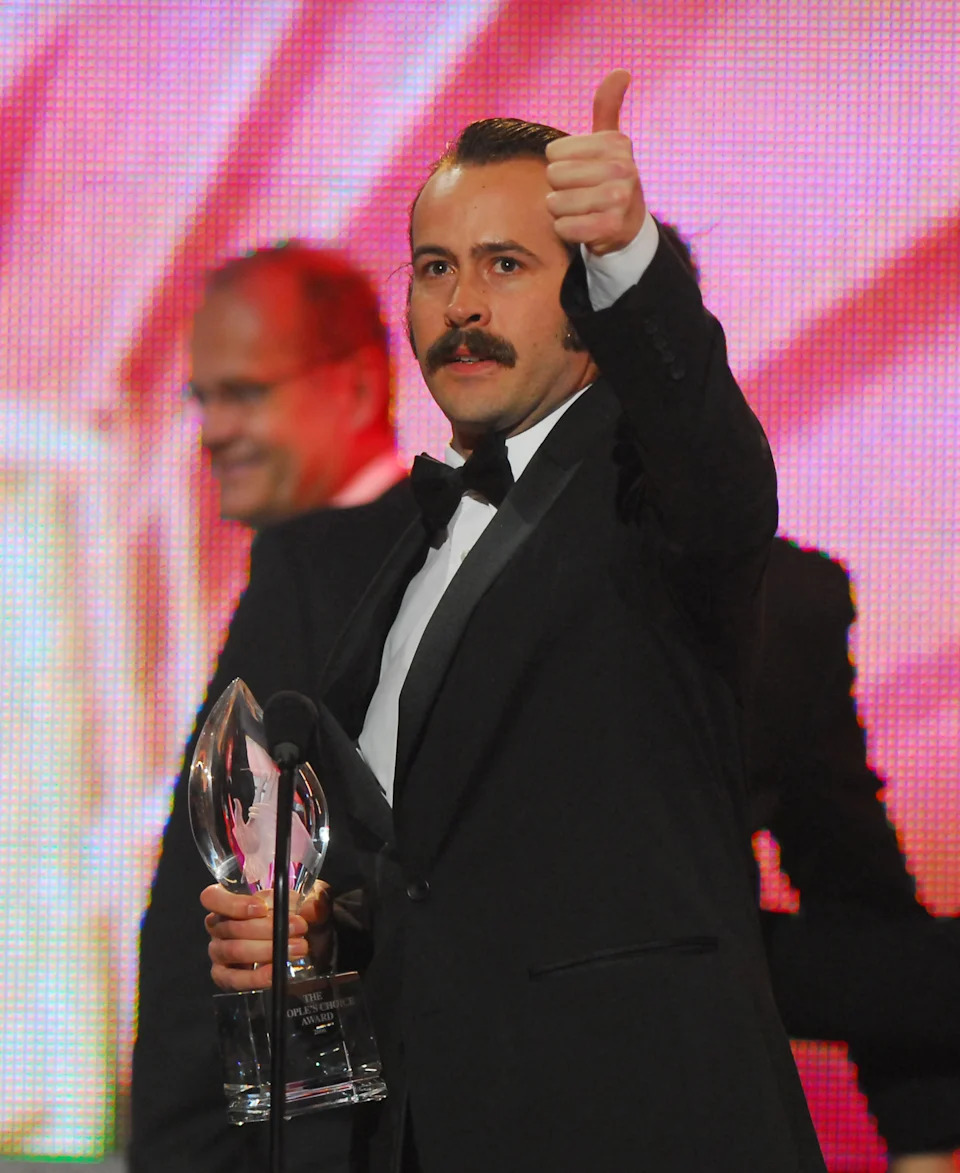 Person in a tuxedo giving a thumbs-up while holding a trophy, standing on stage at an event