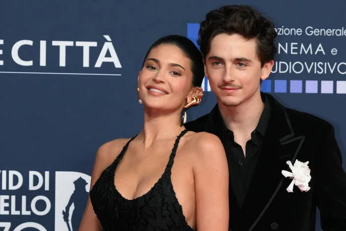 Kylie Jenner and Timothée Chalamet posing on a red carpet. One wears a black gown with a plunging neckline, while the other wears a black suit with a white floral accessory