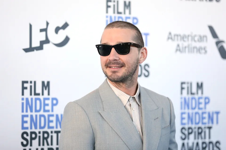 Person in sunglasses and a light-colored suit at the Film Independent Spirit Awards backdrop