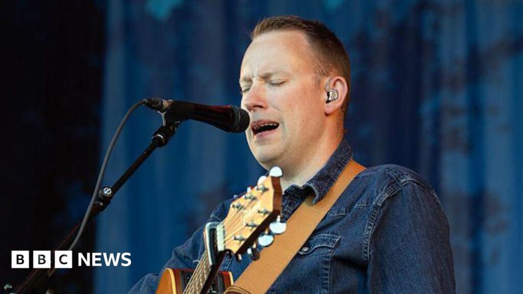 A man standing onstage with an acoustic guitar, singing into a microphone with his eyes closed. He has cropped dark hair and is wearing denim.