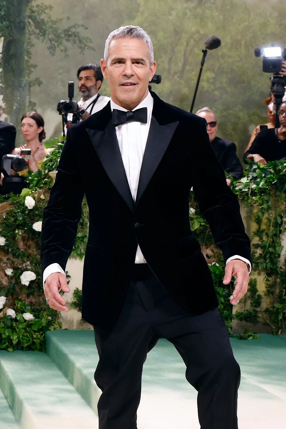 Andy Cohen in a classic tuxedo and bow tie stands on a red carpet as photographers capture the moment