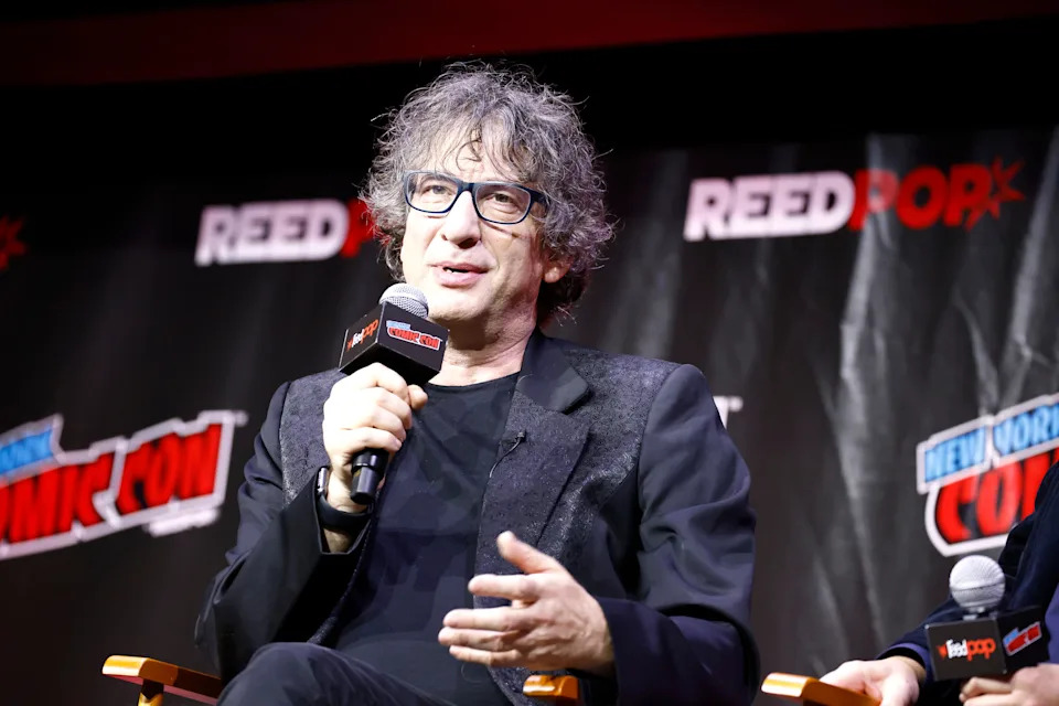 A person on a panel at Comic Con speaks into a microphone, wearing glasses and a casual jacket. Logos are visible in the background