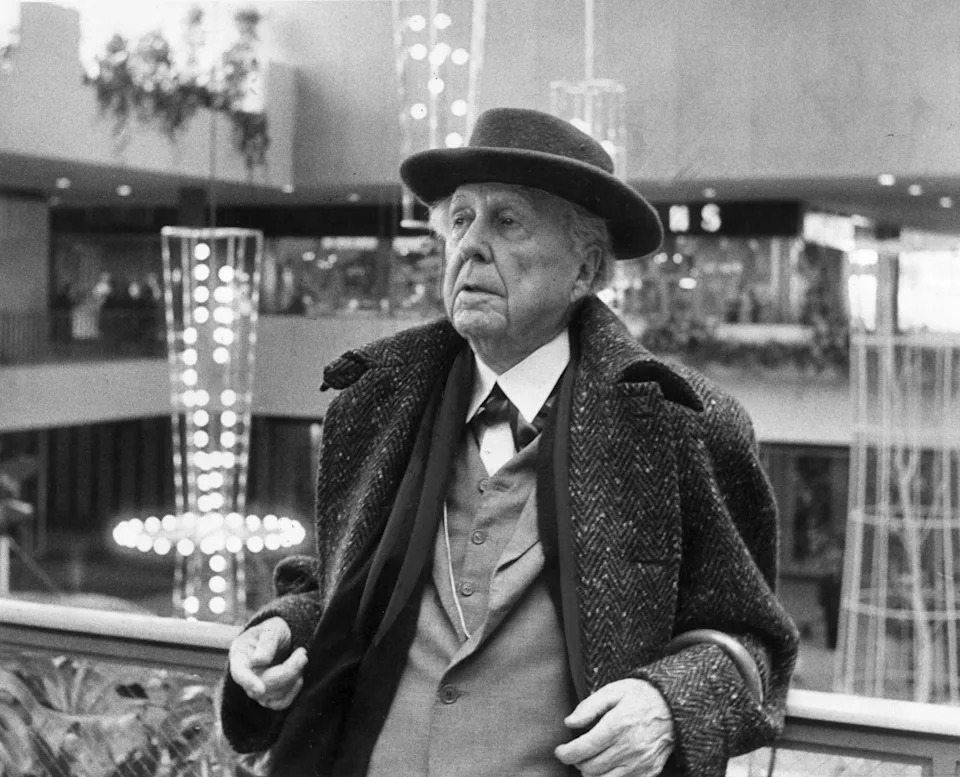 An elderly man in a suit, overcoat, and hat stands in a mall, looking contemplative. The setting includes indoor lighting decorations