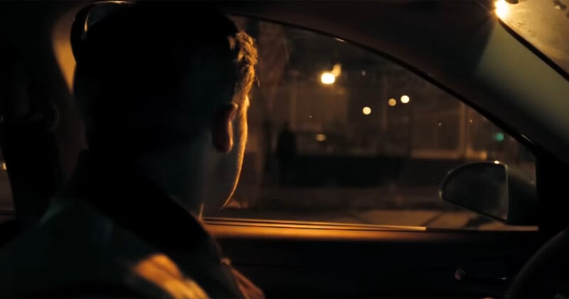 A person sits in a car at night, looking out the passenger-side window toward a dimly lit street with distant streetlights and a shadowy figure on the sidewalk.