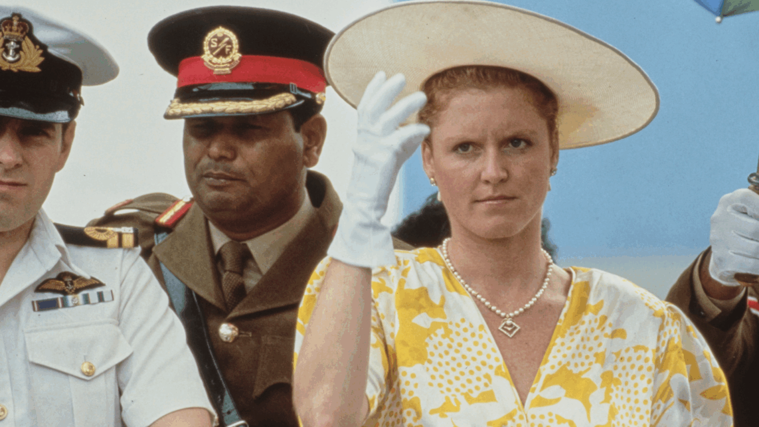 Sarah Ferguson scowling holding on to her yellow hat wearing a pair of white gloves, standing next to Prince Andrew, partially visible