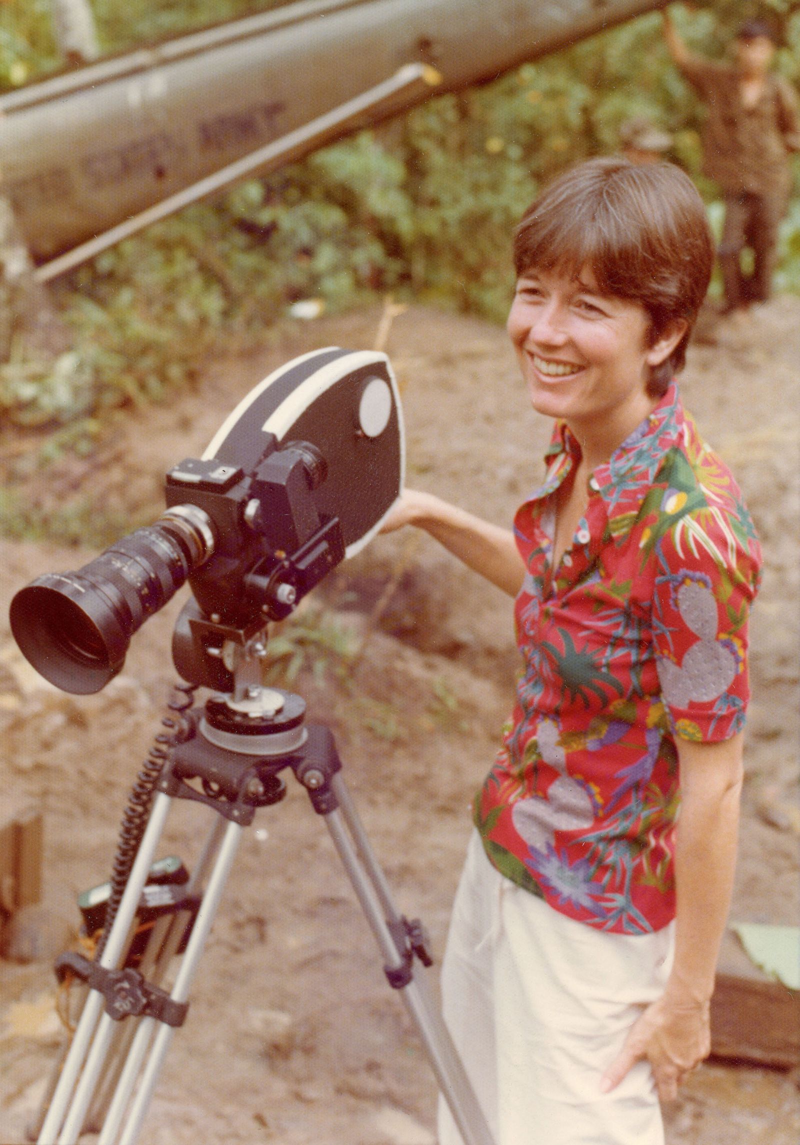 Figure wearing a short sleeve button down stands next to a camera on a tripod smiling off to the distance.