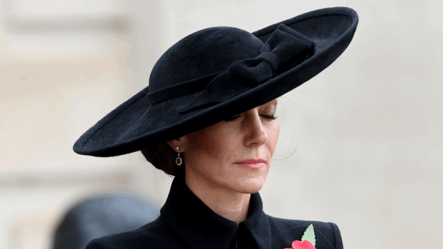 Kate Middleton wearing a black coat and hat closing her eyes