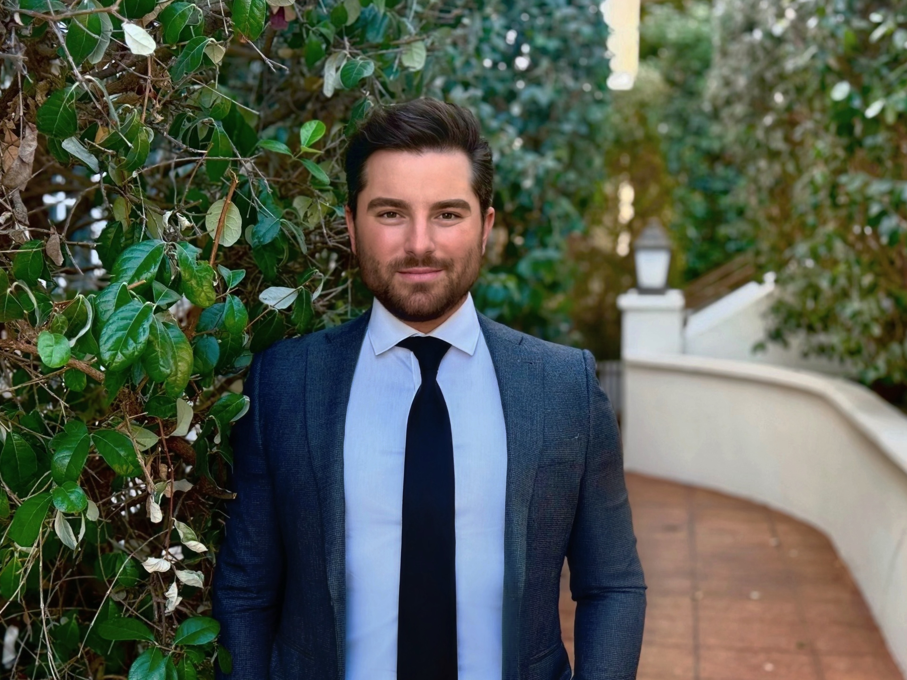 Kyle Boulia wearing a blue suit and tie posing in front of a bush and a stone sidewalk