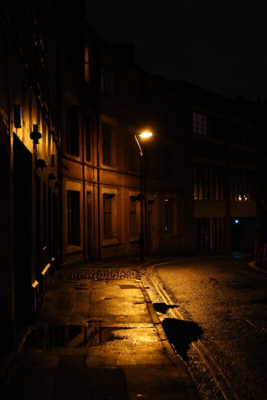 A dimly lit street at night with wet pavement, tall buildings, and a single glowing streetlamp casting light and shadows on the sidewalk and road.