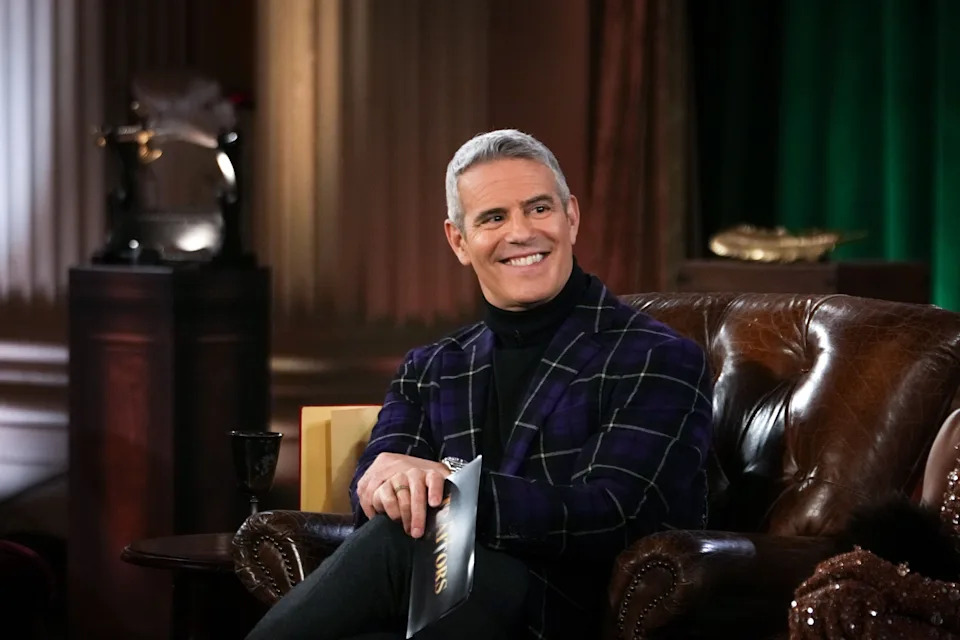 Andy Cohen seated on a leather couch, holding papers and smiling, wearing a plaid blazer over a turtleneck