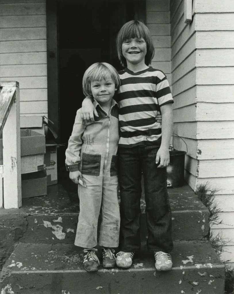 Two children standing on porch steps, one with arm around the other. Both are smiling. One wears overalls, the other wears a striped shirt