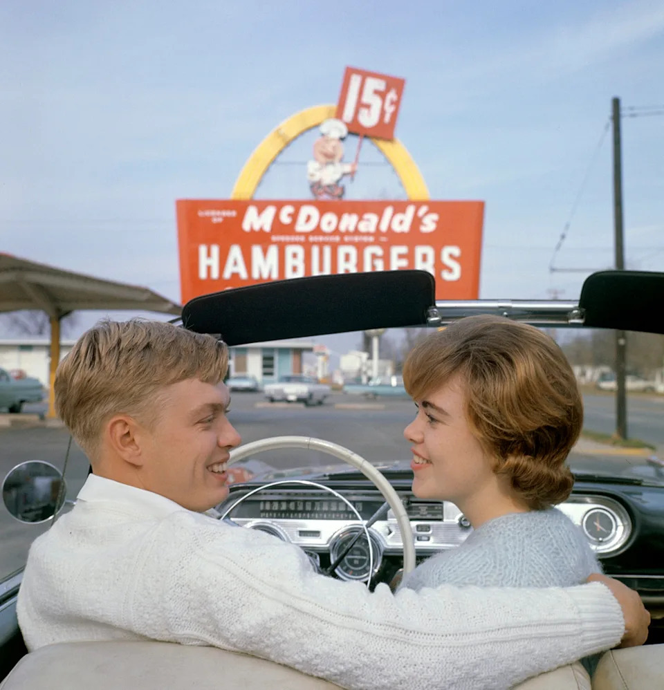 A couple in a vintage convertible smiles at each other in front of a retro McDonald's sign advertising 15-cent hamburgers