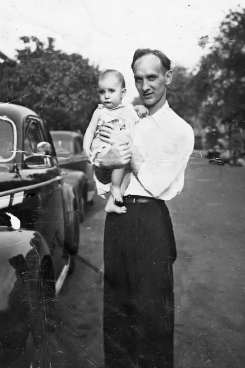 Linda Smith Bianucci Patti as a baby in a dress being held by Grant Smith. He is wearing a white long-sleeved shirt and dark trousers. There are two cars in the background.