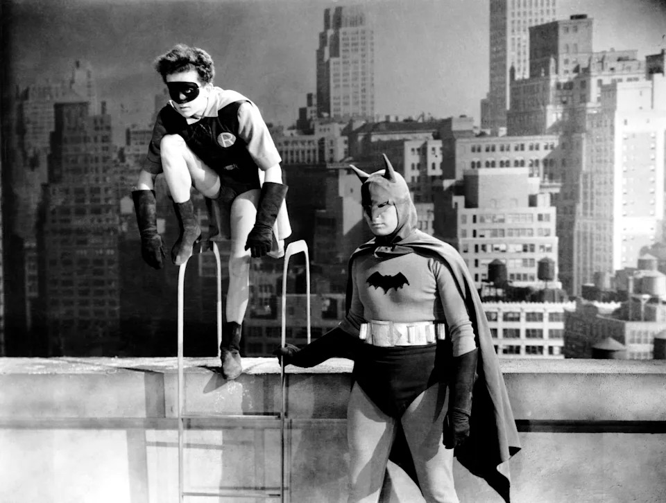 Adam West as Batman and Burt Ward as Robin in costume on a rooftop set with a cityscape background
