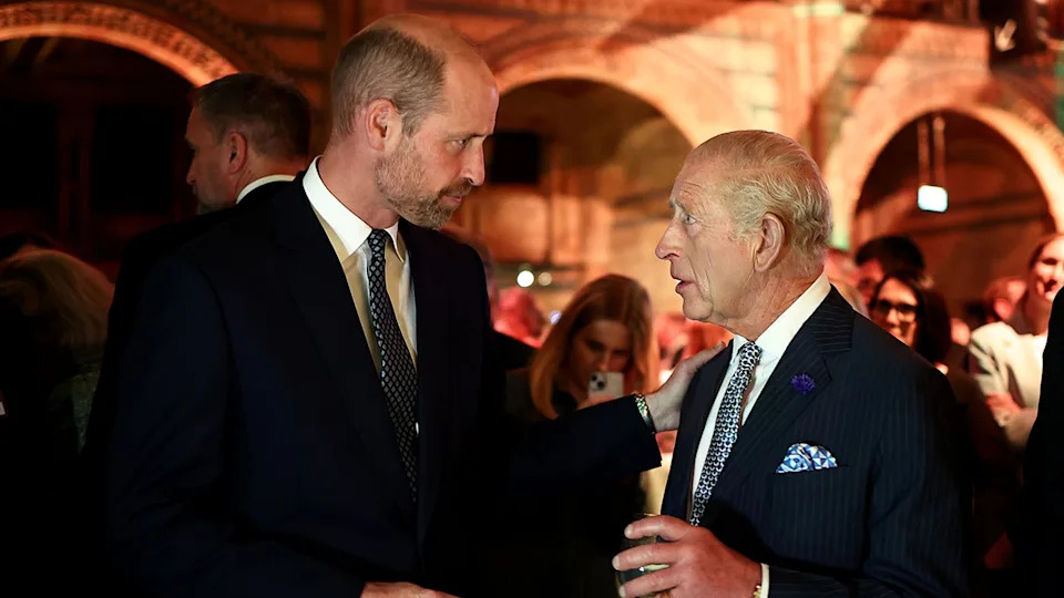 Prince William listening to King Charles speaking during an evening engagement as they both wear matching dark suits.