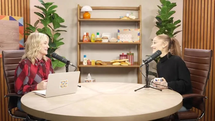 Two people are smiling and talking into microphones in a studio with decor and plants on shelves behind them. They appear engaged in a podcast
