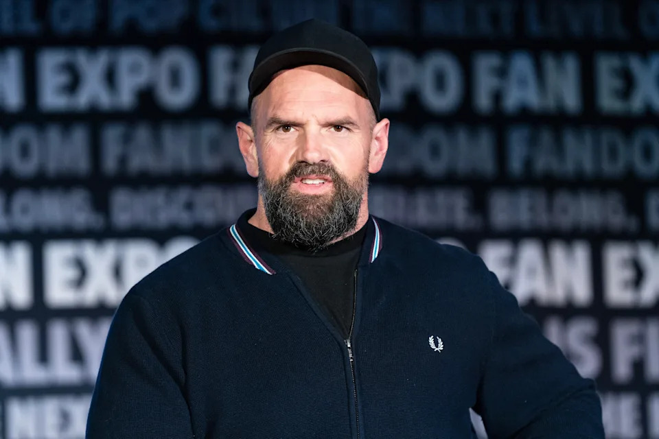 A person wearing a cap and casual sweater stands onstage, appearing to speak at a fan expo event