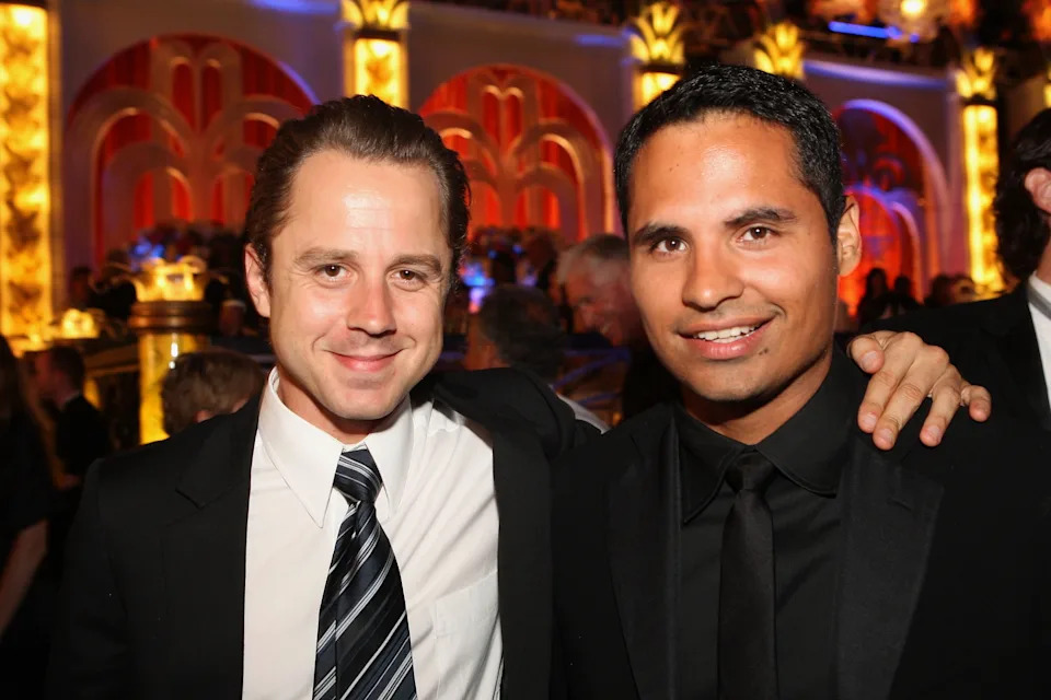 Two men in formal suits with ties, posing together at a glamorous event. One man has his arm around the other, smiling at the camera