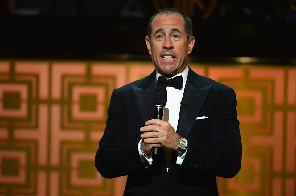 A man in a classic tuxedo speaks on stage, holding a microphone during an event