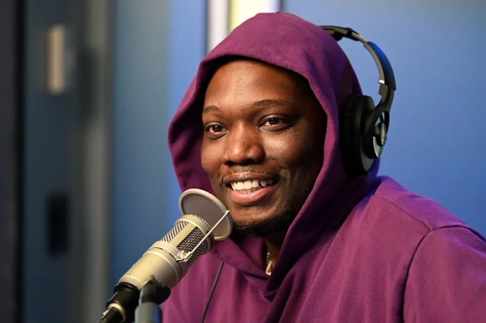 Person in a purple hoodie smiling while speaking into a microphone, wearing headphones in a studio setting