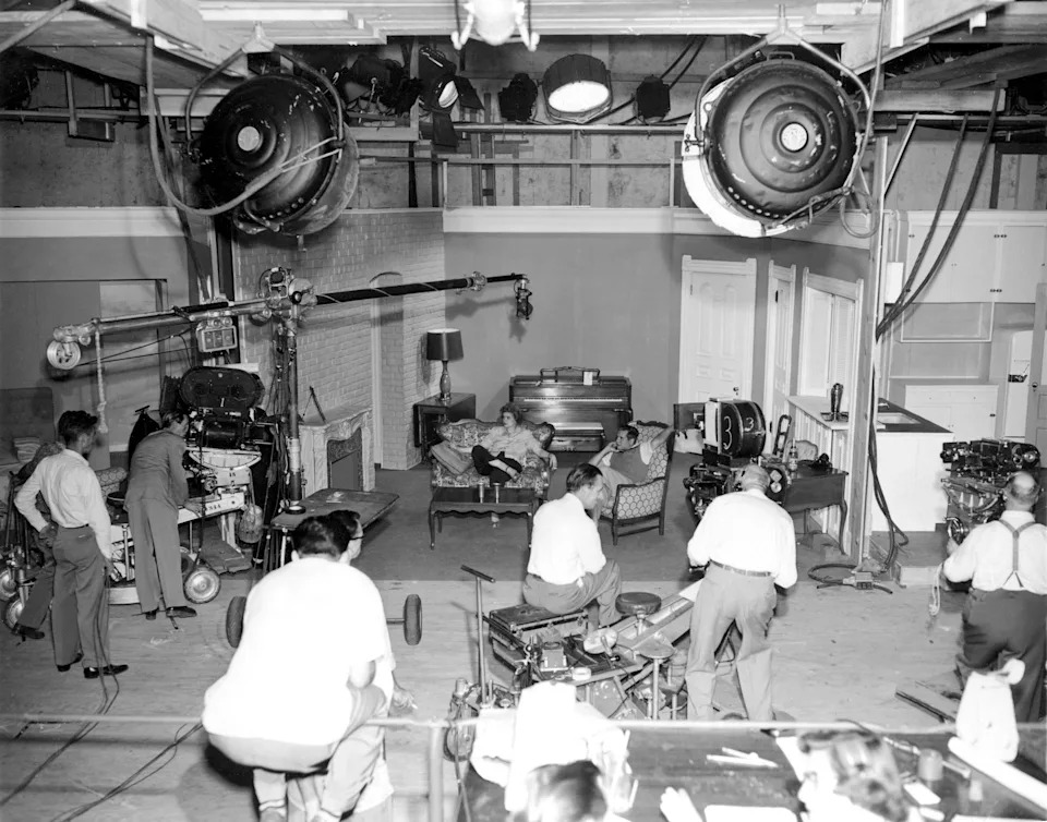 Behind-the-scenes photo of a 1950s TV show set with crew operating cameras and equipment, capturing a living room scene