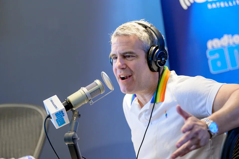 Andy Cohen wearing headphones, sitting at a microphone, gesturing animatedly while speaking in a radio studio