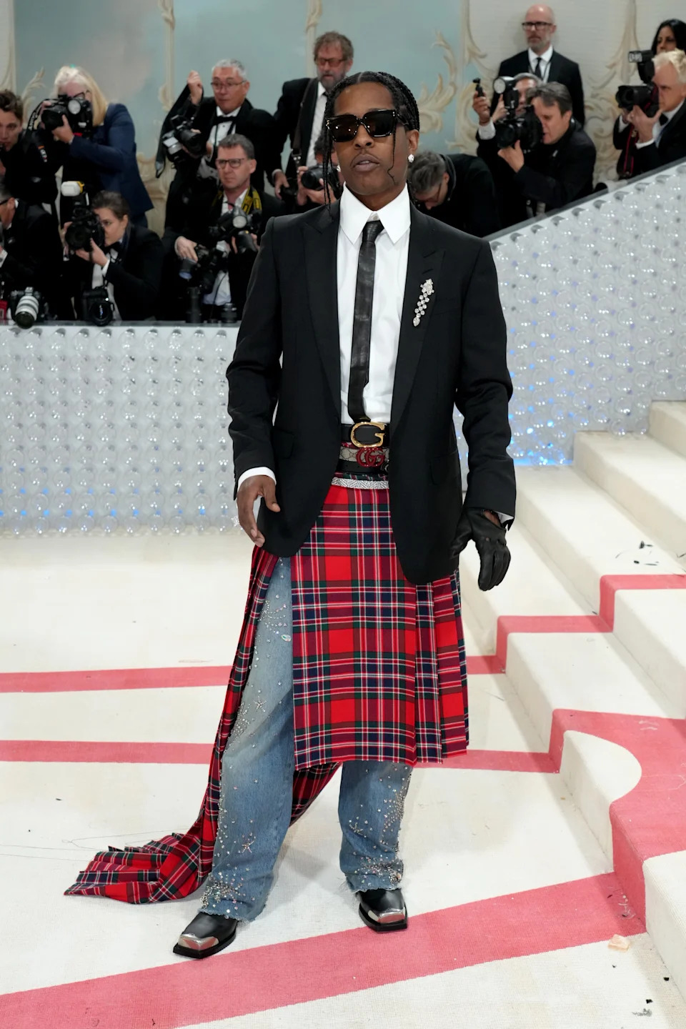A celebrity on a red carpet wears a black blazer, white shirt, tie, and a red tartan skirt over embellished jeans with sunglasses
