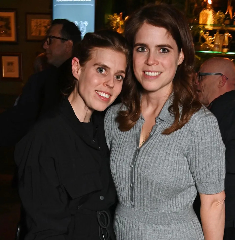 Princess Beatrice wearing black and Princess Eugenie in a gray dress posing in a restaurant.