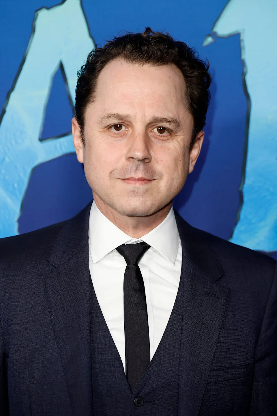 A person in a formal dark suit with a tie stands against a promotional blue background