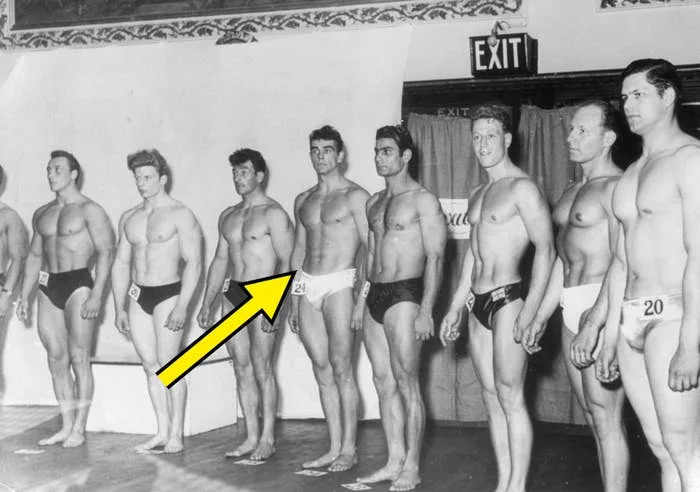 Line-up of male bodybuilders on stage wearing numbered swimwear, likely during a vintage competition