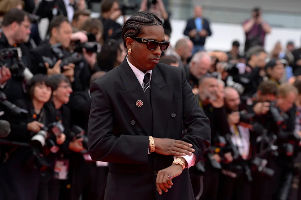 A celebrity on the red carpet wears a sleek black suit with a tie, accessorized with sunglasses and gold jewelry, amidst photographers