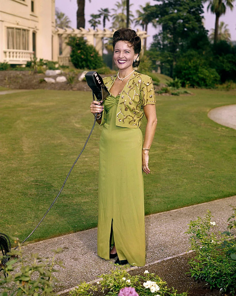 Woman outdoors in an elegant vintage gown with intricate top details, holding a microphone, smiling, with a garden and building in the background