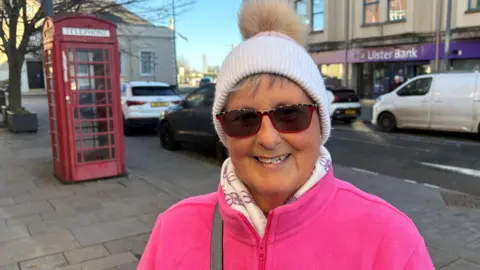 Denis is smiling. She's wearing sunglasses/ The frames have cheeta print at the top and are red at the bottom. She's wearing a oink fleece and a white and pink hat. There's a red phone box behind her in the street.