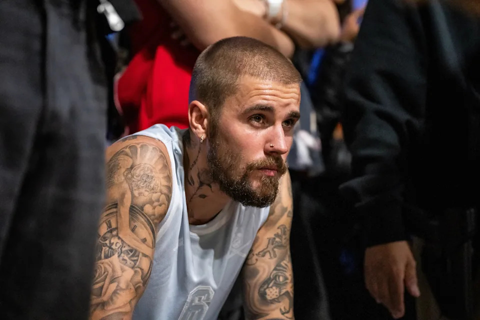 Justin Bieber with tattoos and a trimmed beard wearing a sleeveless shirt leans forward in a crowded room, appearing focused
