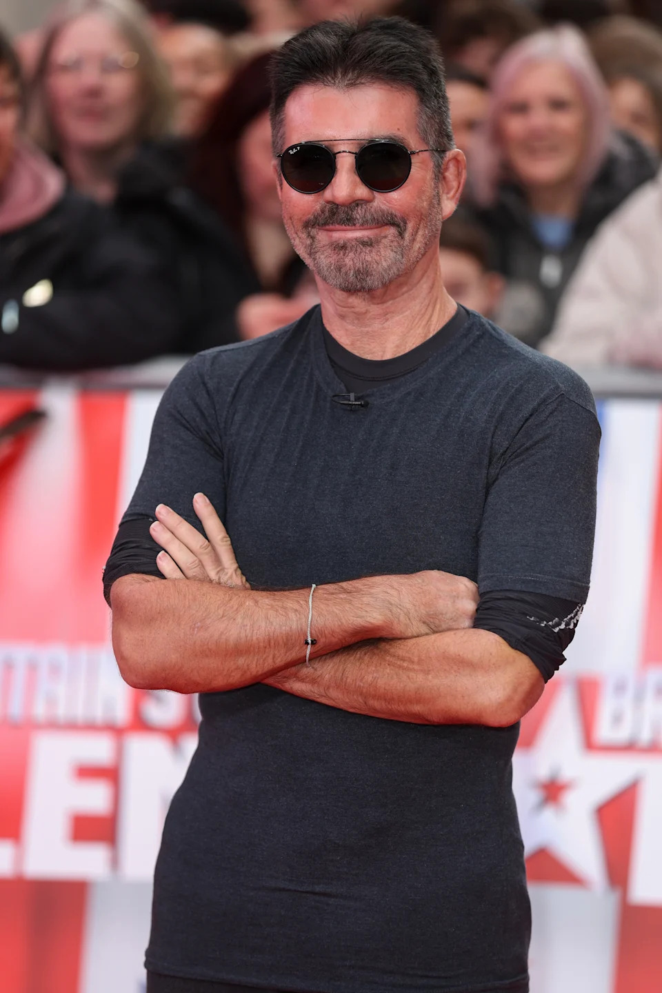 A person in a fitted t-shirt with rolled sleeves, sunglasses, and a bracelet poses confidently on a red carpet, with onlookers in the background