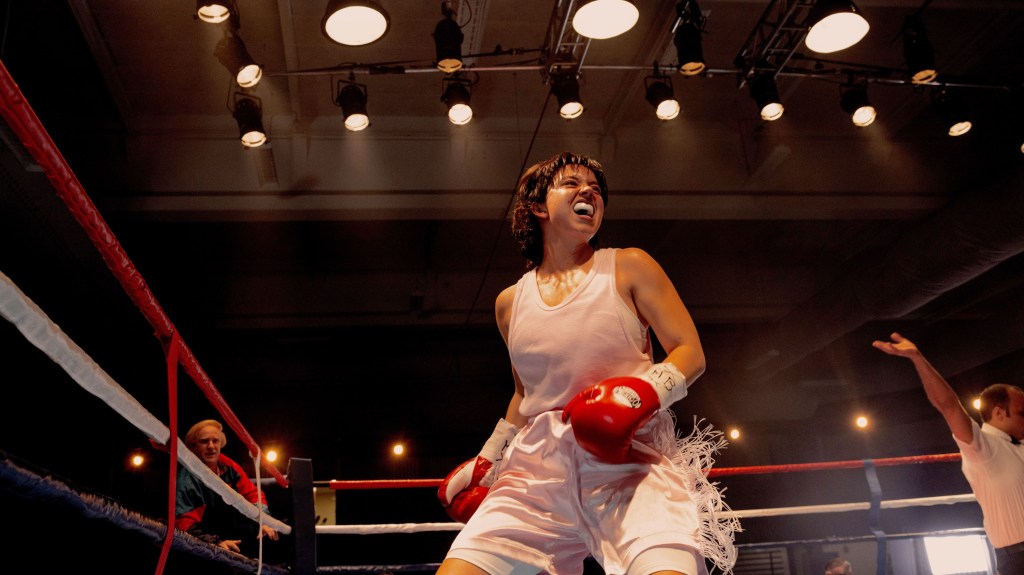 Sydney Sweeney as Christy Martin in a boxing ring, wearing a pink top, white shorts, and red gloves, with Ben Foster as a trainer in the background.