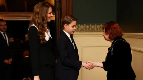 PA Media George and Catherine greet a short woman with red hair as they arrive at the concert hall.