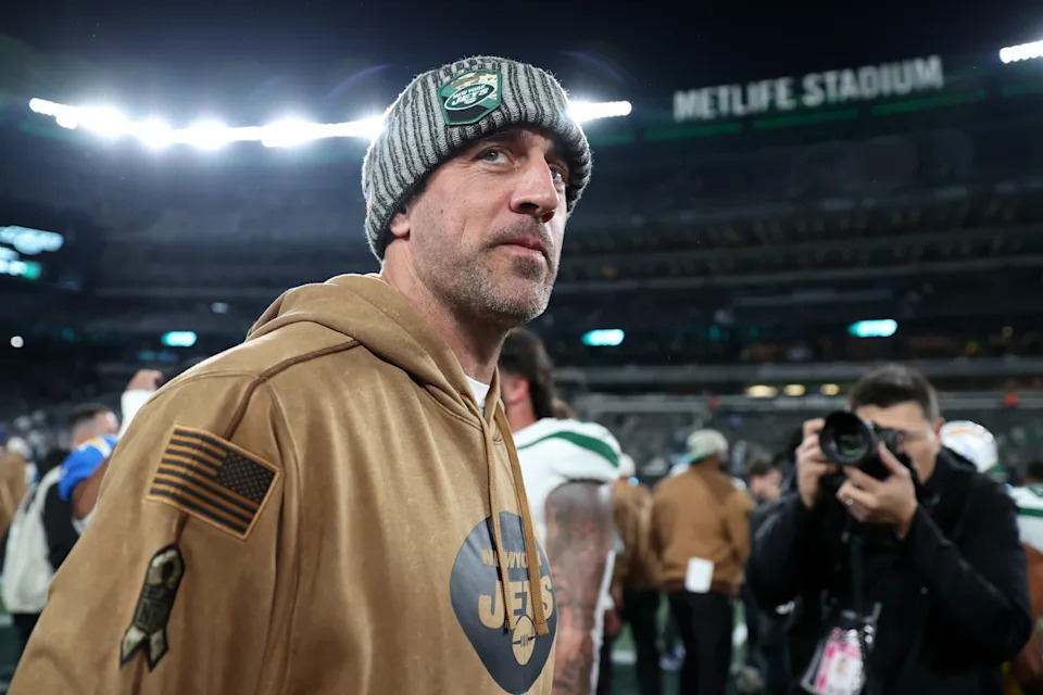 A person in a Jets jacket and beanie stands on a sports field, surrounded by photographers at MetLife Stadium