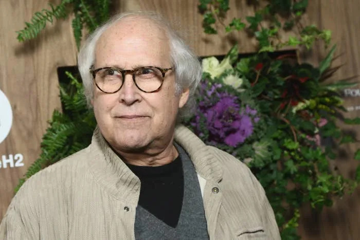 Man in glasses and casual beige jacket standing in front of a floral backdrop at a celebrity event