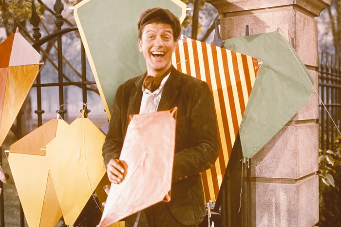 Dick Van Dyke as Bert in Mary Poppins, wearing a brown jacket and hat, smiling broadly and holding a pink kite, with several other kites on the railings behind him.