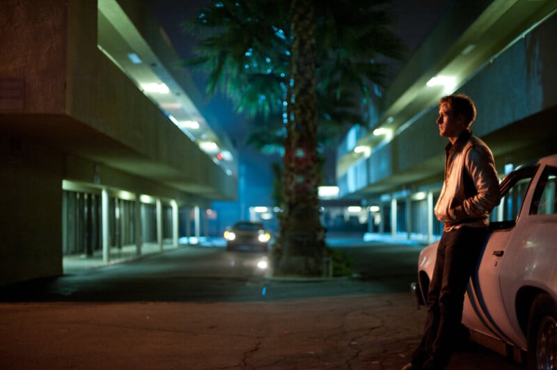 A man in a jacket leans against a white car at night between two dimly lit buildings, with a palm tree and a car with headlights on in the background, creating a moody, cinematic atmosphere.