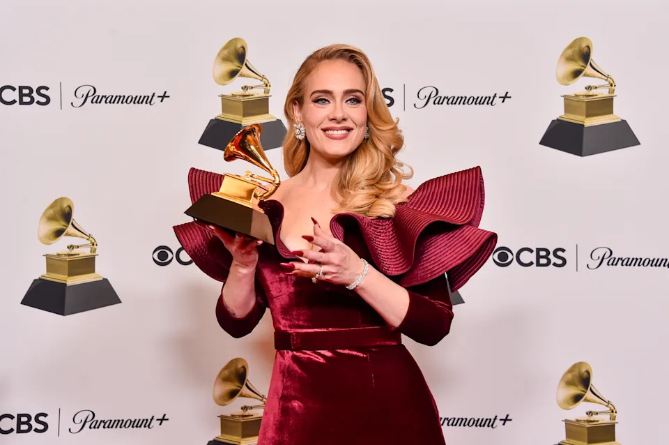 A person in an elegant, ruffled gown holds a Grammy Award on a press stage with logos in the background