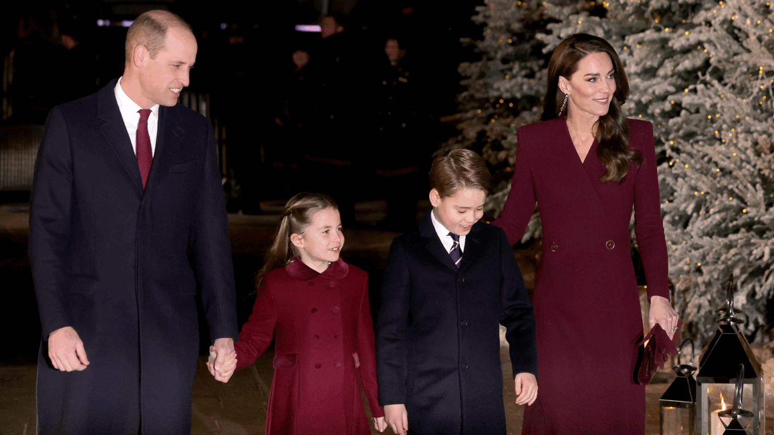Princess Charlotte wearing a burgundy coat and smiling holding Prince William's hand, walking with Prince George and Princess Kate next to a snow covered tree
